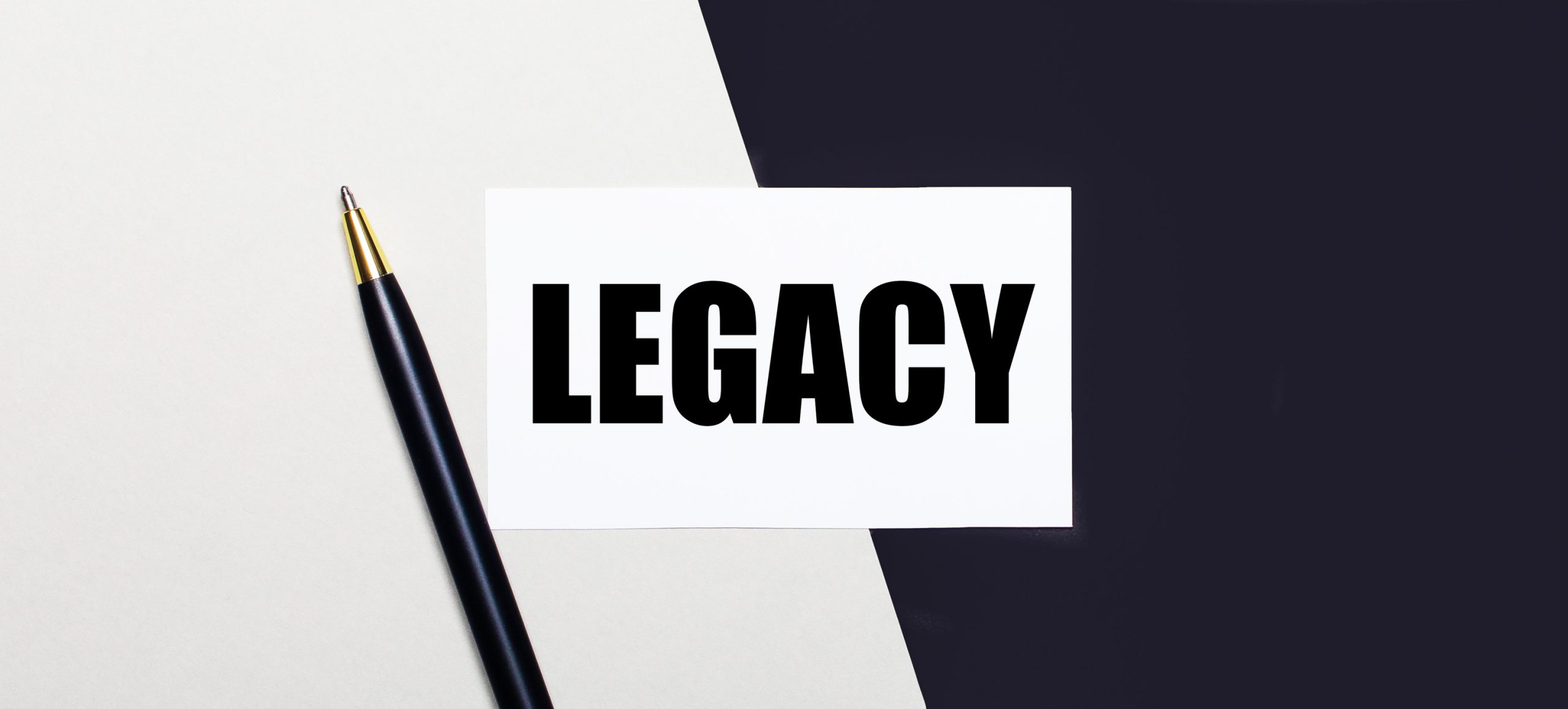 On a black and white background lies a pen and a white card with the text LEGACY